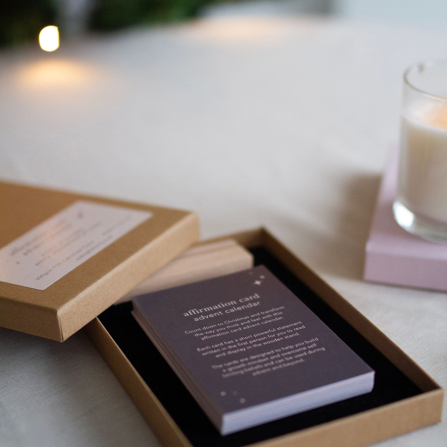 a sparkling Christmas tree in the background, the affirmation card box is open, showing the deck of 24 cards and the wooden display stand